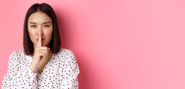 Close-up of mysterious asian woman hiding a secret, hushing and telling to keep quiet, standing over pink background. Copy space