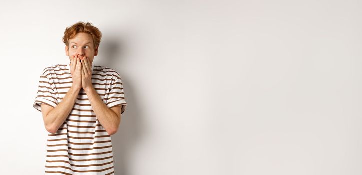Shocked redhead guy gossiping, giggle in hands and looking left impressed, standing over white background.