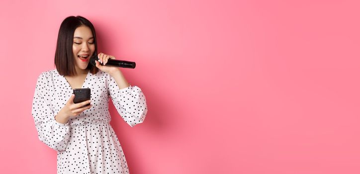 Cute asian woman reading lyrics in smartphone, singing with microphone, standing in trendy dress over pink background.