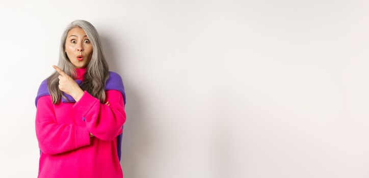 Portrait of impressed asian lady checking out promo offer, pointing at upper left corner and staring at camera amazed, standing in trendy pink sweater over white background.