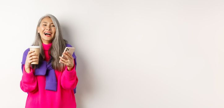 Social networking. Happy asian senior woman drinking coffee and holding smartphone, laughing at camera, standing over white background.