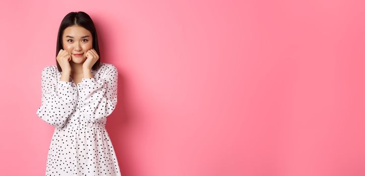 Cute and shy asian girl blushing, touching cheeks and looking at camera silly, standing against pink background.