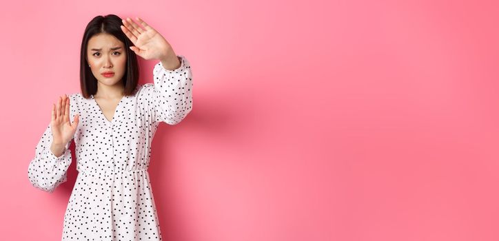 Timid and scared asian woman defending herself, raising arms in protection, victim being attacked, standing over pink background.