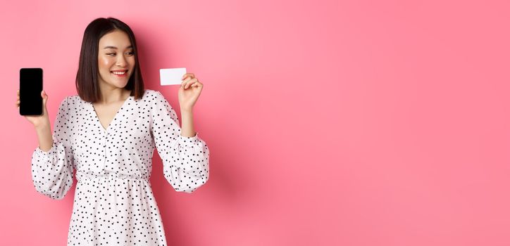 Happy young asian woman internet shopping, showing smartphone screen and looking satisfied at credit card, standing over pink background.