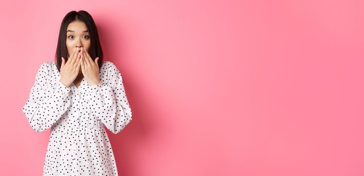 Worried asian girl in dress looking concerned, cover mouth and gasping, staring at camera shocked, standing in cute dress over pink background.