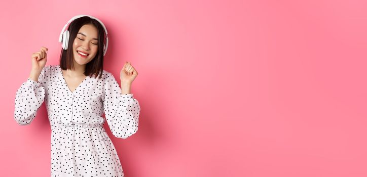 Happy young asian woman dancing and having fun, listening music in headphones, standing over pink background. Copy space