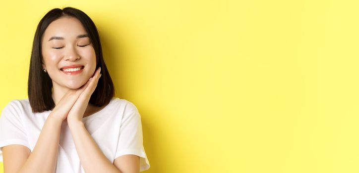Close up of romantic asian woman dreaming of something cute, close eyes and hold hands near face, imaging beautiful thing, standing over yellow background.