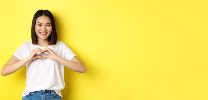 Beautiful asian woman showing I love you heart gesture, smiling at camera, standing against yellow background. Concept of valentines day and romance.