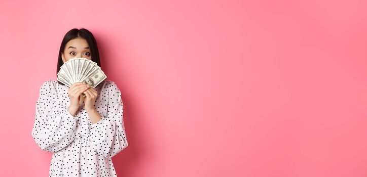 Shopping concept. Cute asian woman hiding face behind money dollars, peeking at camera, standing over pink background.