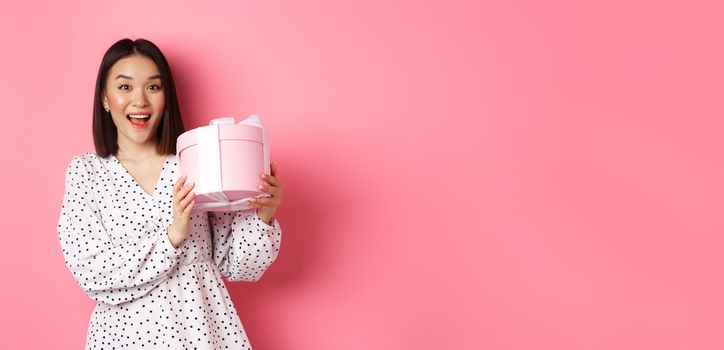 Valentines day. Happy asian woman receiving gift in cute box, smiling excited and thankful, standing in dress against pink background.