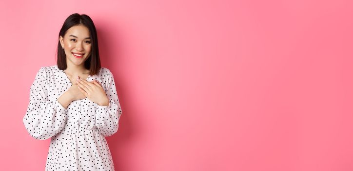 Thankful Korean girl in dress smiling, holding hands on heart and looking grateful at camera, touched with nice gesture, standing over pink background.