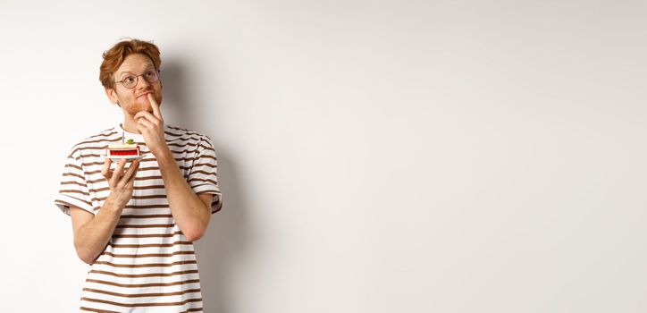 Holidays and celebration concept. Happy young man with red hair and glasses having birthday, holding cake with candle and thinking of b-day wish, standing over white background.