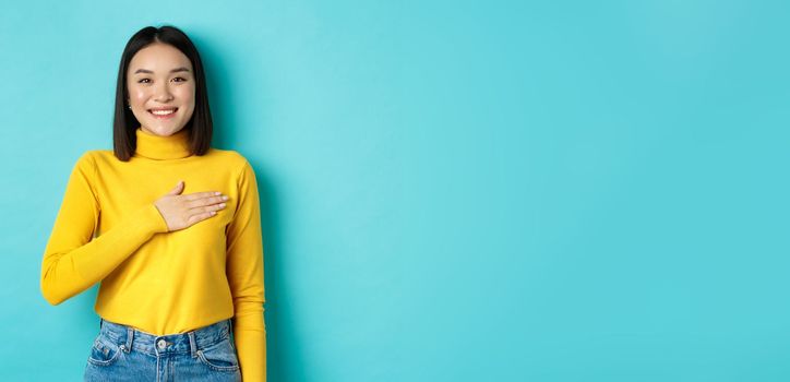 Image of proud smiling asian woman holding hand on heart, showing respect to national anthem, standing over blue background. Copy space