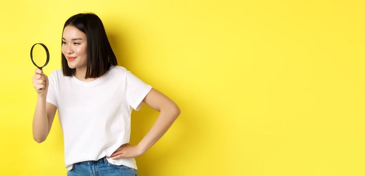 Young asian woman searching for something, looking left through magnifying glass and smiling pleased, investigating, standing over yellow background.