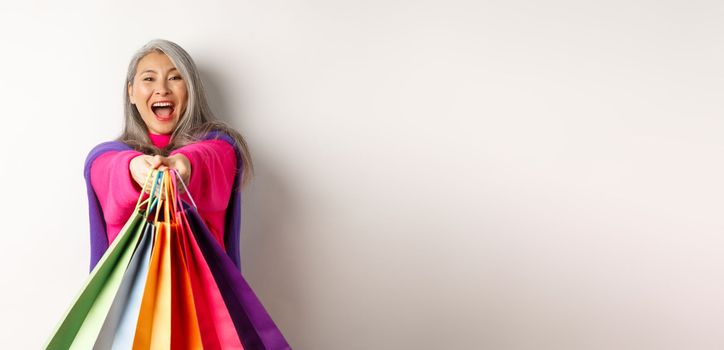 Excited korean lady with grey hair, laughing and showing shopping bags, visit stores with special discounts, standing over white background.