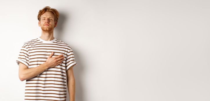 Young bearded redhead man holding hand on heart and looking patriotic, standing during national anthem, standing over white background.