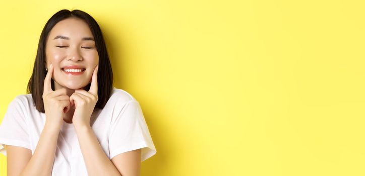 Beauty and skincare. Close up of young asian woman with short dark hair, healthy glowing skin, smiling and touching dimples on cheeks, standing over yellow background.