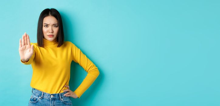 Confident and serious asian woman tell no, showing stop gesture to prohibit and warn, disagree with someone, standing upset against blue background.