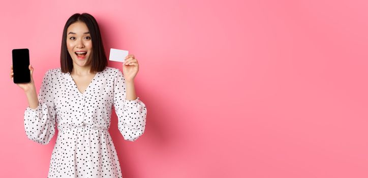 Cute asian woman shopping online, showing bank credit card and mobile screen, smiling and looking at camera, standing over pink background.