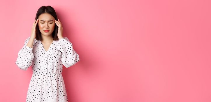 Image of upset asian woman having headache, feeling unwell or dizzy, close eyes and massaging head, suffering migraine, standing over pink background.