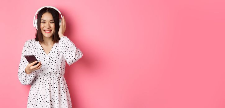 Beautiful asian woman listening music in headphones, using smartphone streaming app and smiling, standing over pink background.