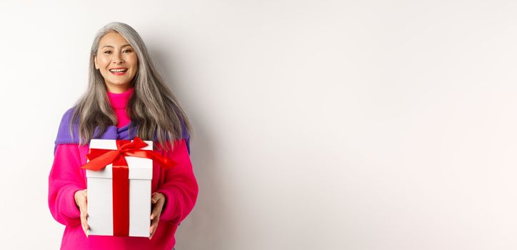 Beautiful asian senior woman smiling, congratulating with valentines day, holding gift in box, standing over white background.