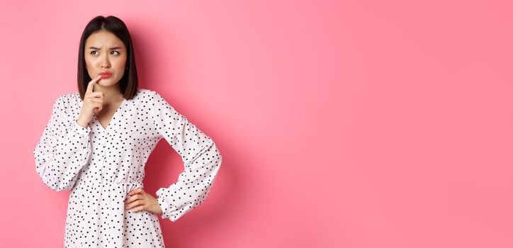 Troubled asian girl making decision, frowning and touching lip while thinking, looking unsure at upper left corner and choosing, standing over pink background.