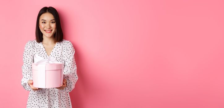 Beautiful asian woman wishing happy holidays, giving you gift in cute box, standing against pink background.