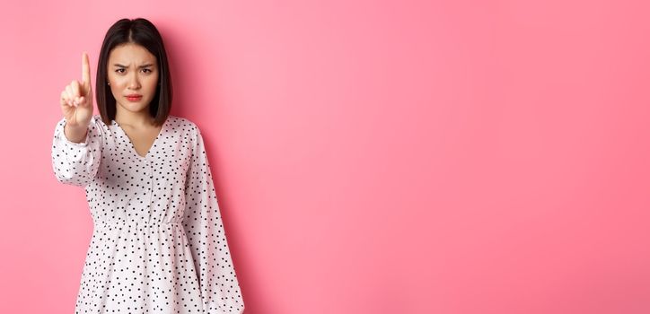 Serious and angry asian woman telling to stop, frowning and showing finger in disapproval, prohibit something, standing in dress against pink background.