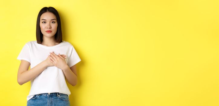 Beauty and fashion concept. Beautiful asian woman holding hands on heart and looking thoughtful at camera, keeping memories in soul, standing over yellow background.