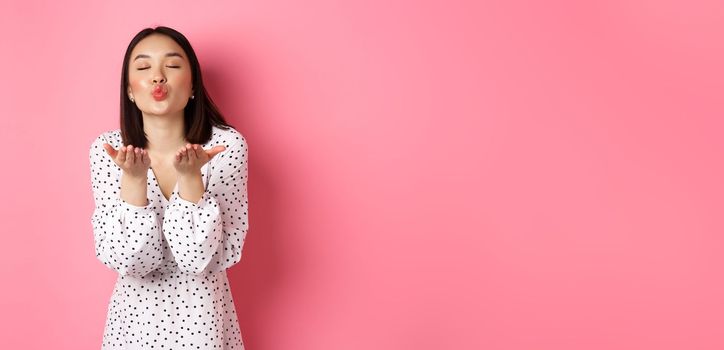 Beautiful chinese girl in dress blowing air kisses, sending mwah at camera, standing over pink background.