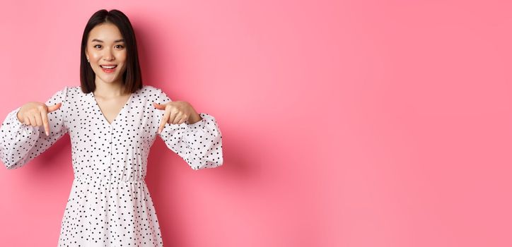 Look there. Cute asian woman in dress, pointing fingers down at copy space, showing product discount and smiling, standing over pink background.