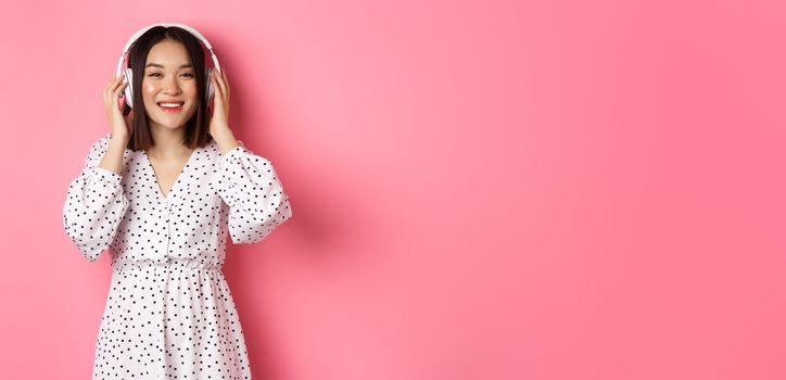 Romantic asian woman smiling happy, listening music in headphones and looking at camera, standing over pink background.