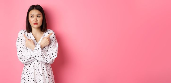 Concerned asian woman having doubts, pointing sideways and looking left with hesitant and sad face, need help with choice, standing over pink background.