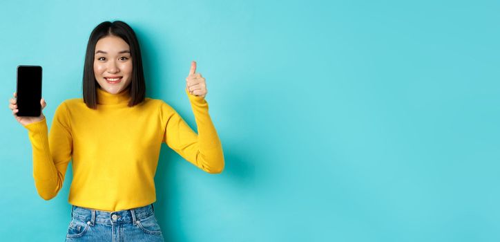 Technology and people concept. Cheerful asian girl in yellow sweater showing blank smartphone screen and thumbs up, demonstrate online offer, standing over blue background.