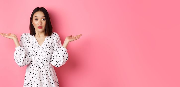 Confused and clueless asian girl shrugging, staring at camera puzzled, dont know what to do, spread hands sideways, standing over pink background.