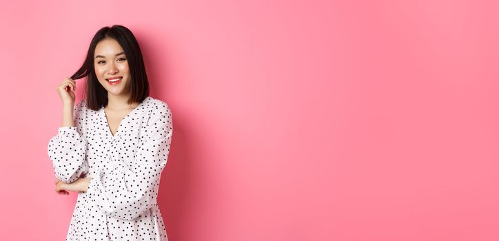 Cute asian female brunette in trendy dress, playing with hair and smiling flirty, standing in dress over pink background.