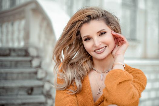 A middle-aged woman looks like a good blonde with curly beautiful hair and makeup on the background of the building. She is wearing a yellow sweater and looking at the camera