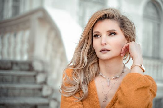 A middle-aged woman looks like a good blonde with curly beautiful hair and makeup on the background of the building. She is wearing a yellow sweater