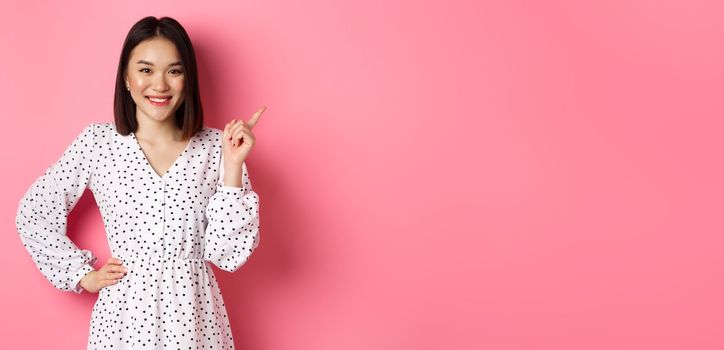 Beautiful asian female model smiling, pointing finger at upper right corner copy space, showing advertisement banner, standing over pink background.