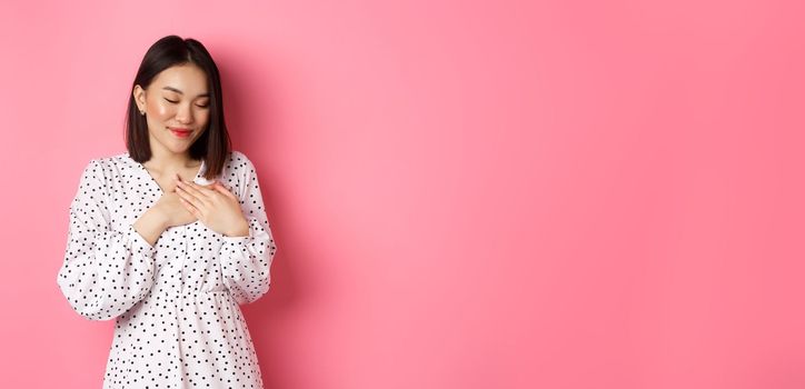 Beautiful korean girl in trendy spring dress dreaming, holding hands on heart, smiling with closed eyes, imaging something or having heartwarming memory, pink background.