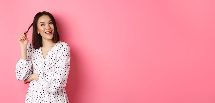 Dreamy cute asian girl thinking, imaging shopping and smiling, looking upper left corner with thoughtful smile, standing in dress over pink background.