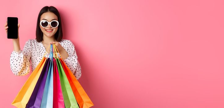 Attractive asian woman showing smartphone app and shopping bags, buying online via application, standing over pink background.