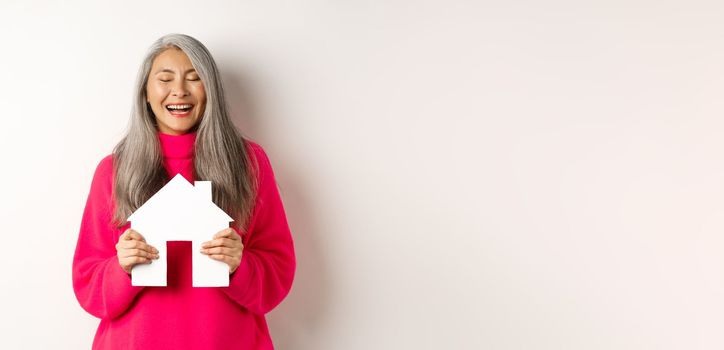 Real estate. Happy asian grandmother laughing with eyes closed, holding paper house model, dreaming about paper house model, standing over white background. Copy space