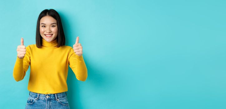 Cheerful asian female model showing thumbs up gesture, smiling and looking impressed, praise good product, standing over blue background.