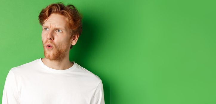 Confused redhead man staring left and looking worried, standing against green background.