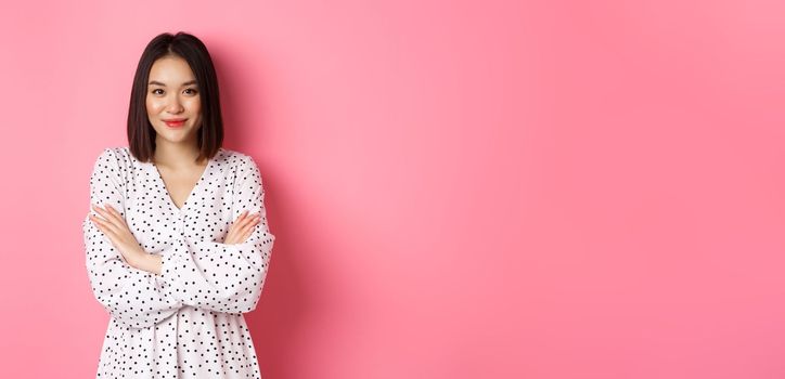 Beautiful asian female model standing in dress, cross arms on chest and smiling at camera, standing over pink background.