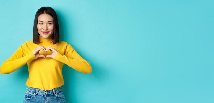 Attractive japanese girl in yellow sweater, showing heart gesture and say I love you, looking heartfelt at camera, standing over blue background.