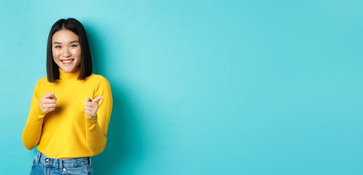 Smiling young asian woman pointing fingers at camera, choosing you, inviting to event, standing happy against blue background.