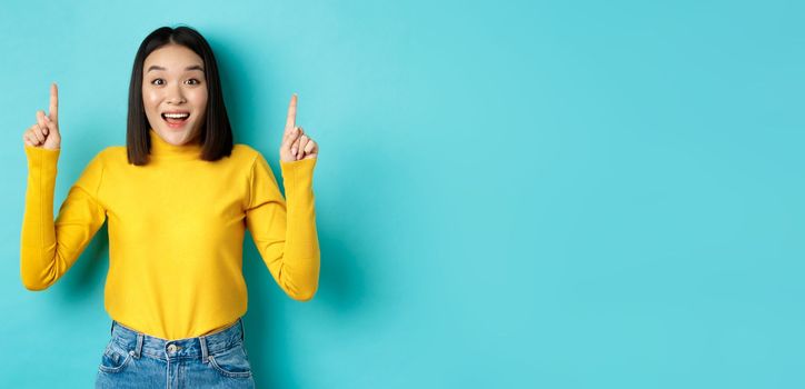 Shopping concept. Impressed young korean woman showing advertisement on top, pointing fingers up and gasping amazed, saying wow, standing over blue background.